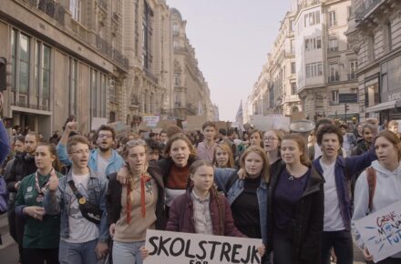 Duża grupa osób idących ulicą w ramach protestu klimatycznego. Na czele idzie Greta Thunberg z transparentem w języku szwedzkim.